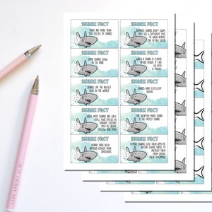 May include: A stack of shark fact cards with cartoon shark illustrations and facts. The cards have a light blue and white background. Two pink pens are in the image, one with the text "IS IT FRIDAY YET?"