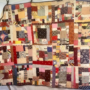 Puede incluir: Un edredón de retazos con una variedad de patrones y colores de tela, incluyendo rojo, rosa, azul y marrón. El edredón está hecho de muchos trozos de tela pequeños e irregulares cosidos juntos. El edredón está colgado en una pared.