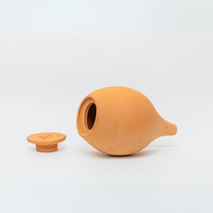May include: A terracotta ceramic jar with a lid. The jar is a light orange color and has a rounded body with a long, thin spout. The lid is a small, round disc with a raised rim.