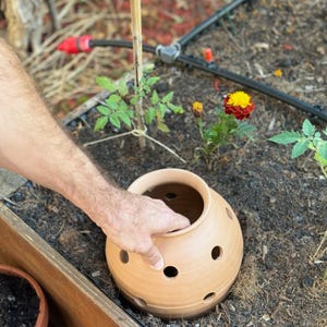 Op de afbeelding: Een terracotta pot met drainagegaten wordt in een tuinbed geplaatst. De pot wordt vastgehouden door een hand. Het tuinbed is gevuld met bruine aarde en heeft een zwart irrigatiesysteem.