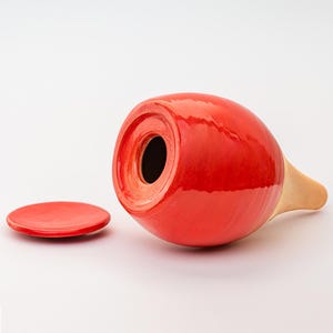 Könnte beinhalten: Eine rote dekorative Keramikvase mit passendem Deckel. Die Vase hat einen abgerundeten Körper und einen hellbraunen, sich verjüngenden Ausguss. Der Deckel ist ein kleiner, flacher Kreis. Die Vase liegt auf der Seite.