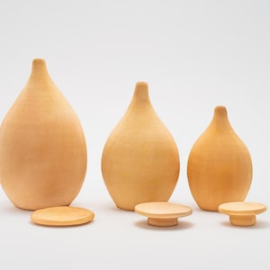 May include: Three wooden vases in different sizes, all with a light brown color and a rounded, teardrop shape. The vases are arranged in a row from largest to smallest, with the largest vase on the left and the smallest vase on the right. Each vase is sitting on a small, round wooden base.