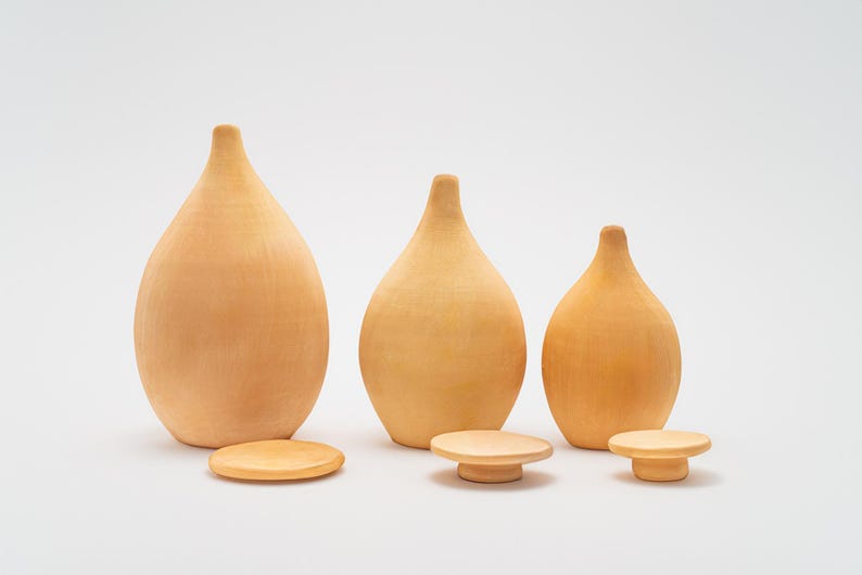 May include: Three wooden vases in different sizes, all with a light brown color and a rounded, teardrop shape. The vases are arranged in a row from largest to smallest, with the largest vase on the left and the smallest vase on the right. Each vase is sitting on a small, round wooden base.