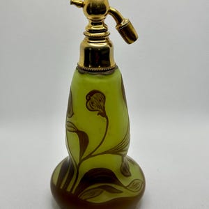 Puede incluir: Un frasco de perfume de vidrio verde con un atomizador dorado y un diseño floral. El frasco tiene una base bulbosa y un cuello delgado.
