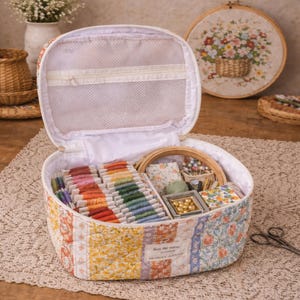 May include: Open sewing kit filled with colourful threads, needles, and other supplies. The kit has floral and patchwork patterns. A floral embroidery hoop is in the background, along with a small basket and scissors.
