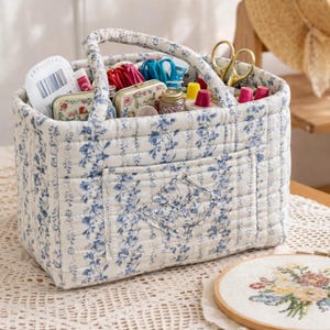 May include: A fabric sewing basket with a blue floral design, containing sewing supplies. The basket has a handle and a front pocket. Inside are spools of thread, scissors, and other craft items. An embroidery hoop with cross-stitch is in the foreground.