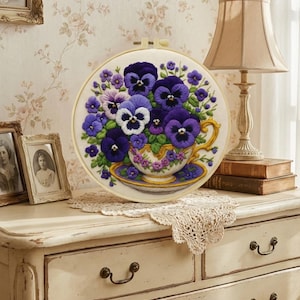 May include: A round embroidery hoop featuring a floral design. The artwork showcases a teacup overflowing with purple and white pansies. The teacup and saucer are detailed with gold accents, set against a cream-colored background.