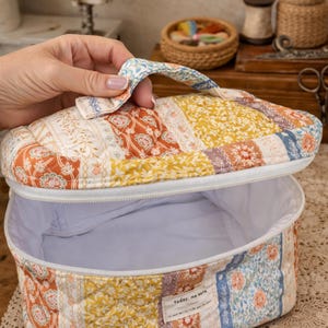 以下が含まれることがあります： パッチワークスタイルのキルトソーイングボックス。持ち手付きで、開くと白い内側が見えます。オレンジ、黄色、青、白の色合いで、さまざまな花柄と幾何学模様が特徴です。「today, me ears」の文字が小さなラベルに表示されています。