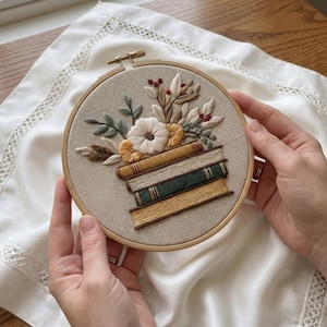 May include: Embroidered hoop art depicting a stack of books topped with a floral arrangement. The embroidery features a white flower, yellow accents, and green and brown leaves. The hoop is held by hands.