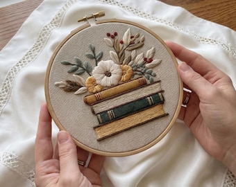 May include: An embroidered hoop art piece featuring a stack of books with a floral arrangement on top. The embroidery includes a white flower, yellow accents, and green and brown leaves. The hoop is held by hands.