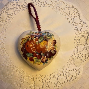 May include: Heart-shaped ornament with a fruit and floral design. The ornament features a burgundy ribbon for hanging. The design includes oranges, grapes, and berries. The ornament is placed on a white doily.