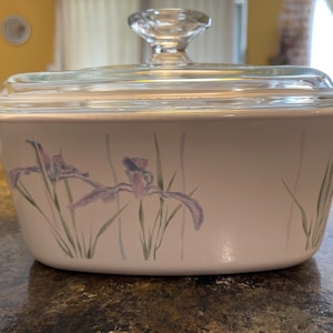 May include: A white casserole dish with a clear glass lid and a clear glass knob. The dish features a floral design of purple irises and green stems. The dish has two small handles on either side.