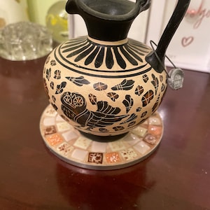 May include: A decorative ceramic vase with a black handle and spout, featuring a black and beige design with an owl motif. The vase sits on a mosaic tile coaster with a silver rim. The vase is a unique home decor item.