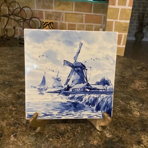 May include: Square ceramic tile featuring a blue and white Dutch windmill scene. The artwork depicts windmills, sailboats, and water, with a cloudy sky background. The tile is displayed on a brass stand.