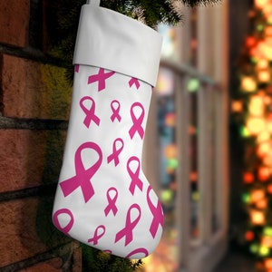 Op de afbeelding: Witte kerstsok met een patroon van roze bewustzijns linten. De sok heeft een witte manchet en hangt aan een bakstenen muur. De achtergrond is vervaagd met kerstverlichting.