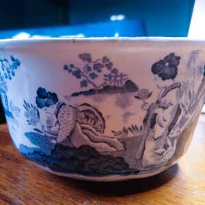 Puede incluir: Un tazón de cerámica blanco con un patrón floral azul y blanco. El tazón presenta una escena con una figura con ropa tradicional y un paisaje con árboles y un puente.