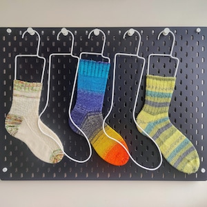 May include: Three knitted socks hanging on white wire hangers against a black pegboard. The left sock is cream with multicoloured speckles. The middle sock features a blue, purple, and orange gradient. The right sock has green and grey stripes.