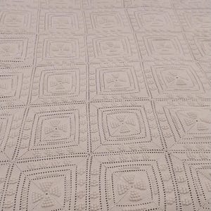 May include: Cream-colored, crocheted blanket with a repeating square pattern. The blanket features a detailed, textured design with a scalloped edge. Two pink pillows are visible in the background.
