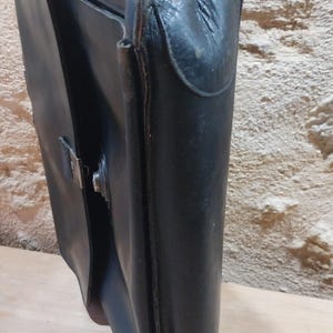 Puede incluir: Un malet&iacute;n de cuero negro con un cierre met&aacute;lico. El malet&iacute;n est&aacute; abierto, revelando el interior. El cuero parece desgastado, lo que sugiere edad y uso. La bolsa est&aacute; dise&ntilde;ada para transportar documentos y otros elementos esenciales.