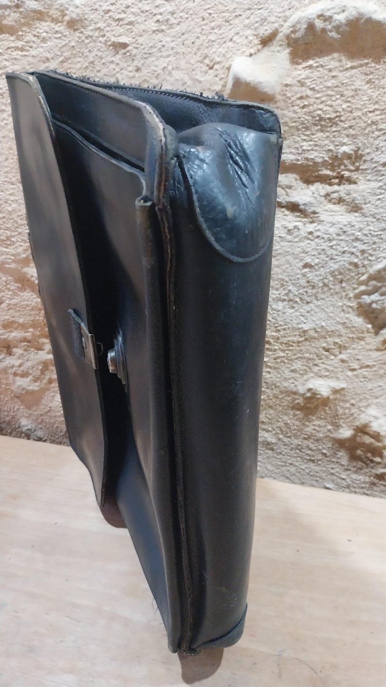 Puede incluir: Un malet&iacute;n de cuero negro con un cierre met&aacute;lico. El malet&iacute;n est&aacute; abierto, revelando el interior. El cuero parece desgastado, lo que sugiere edad y uso. La bolsa est&aacute; dise&ntilde;ada para transportar documentos y otros elementos esenciales.