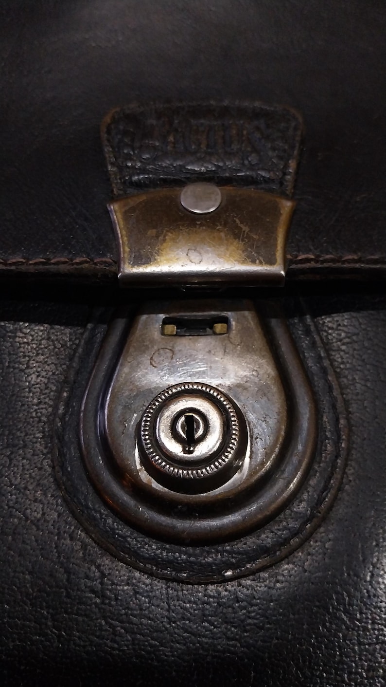 Puede incluir: Primer plano de un cierre de bolso de cuero negro. El cierre es de metal deslustrado con un dise&ntilde;o de ojo de cerradura. El cuero tiene un aspecto texturizado y el cierre met&aacute;lico muestra signos de desgaste.