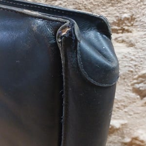 Puede incluir: Primer plano de un bolso de cuero negro, que muestra la esquina y el asa. El cuero parece desgastado con algunos da&ntilde;os en los bordes. El fondo es una superficie texturizada de color marr&oacute;n claro.
