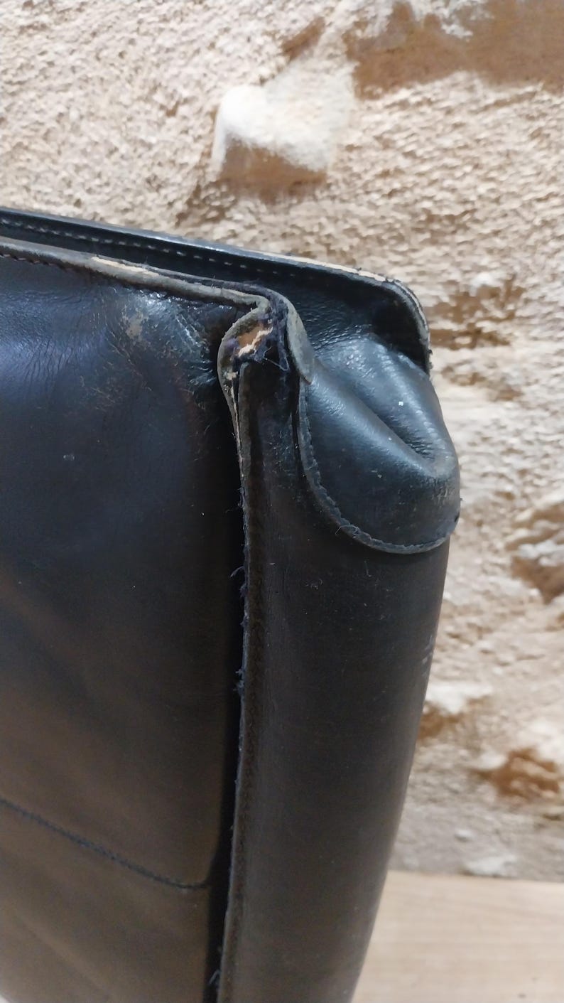 Puede incluir: Primer plano de un bolso de cuero negro, que muestra la esquina y el asa. El cuero parece desgastado con algunos da&ntilde;os en los bordes. El fondo es una superficie texturizada de color marr&oacute;n claro.