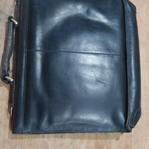 Puede incluir: Un malet&iacute;n de cuero negro con asa y herrajes met&aacute;licos. La bolsa rectangular tiene un aspecto ligeramente desgastado, con pliegues visibles y un brillo sutil. El dise&ntilde;o incluye una costura central y esquinas reforzadas.