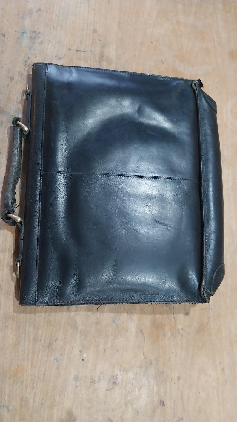 Puede incluir: Un malet&iacute;n de cuero negro con asa y herrajes met&aacute;licos. La bolsa rectangular tiene un aspecto ligeramente desgastado, con pliegues visibles y un brillo sutil. El dise&ntilde;o incluye una costura central y esquinas reforzadas.