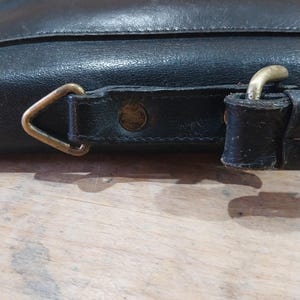Puede incluir: Primer plano de un bolso de cuero negro con una hebilla de lat&oacute;n y una anilla en D. El bolso tiene una superficie texturizada y un borde cosido. Los herrajes son de color dorado brillante.