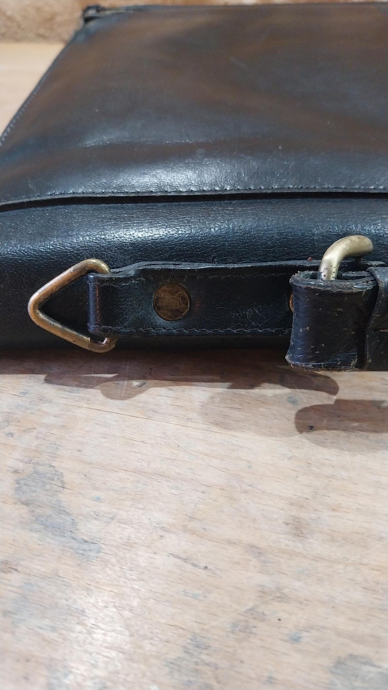 Puede incluir: Primer plano de un bolso de cuero negro con una hebilla de lat&oacute;n y una anilla en D. El bolso tiene una superficie texturizada y un borde cosido. Los herrajes son de color dorado brillante.