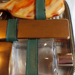 Puede incluir: Un neceser de viaje vintage de cuero con cierre de cremallera. El neceser contiene un peine, un cepillo, una botella de colonia y un pequeño recipiente de metal. El neceser está forrado con tela verde y tiene detalles dorados.