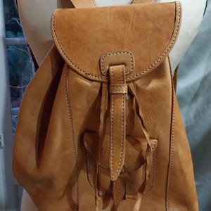Op de afbeelding: Een bruinleren rugzak met verstelbare bandjes en een klepsluiting. De tas heeft twee voorzakken en decoratieve stiksels. De rugzak wordt op een mannequin gepresenteerd.