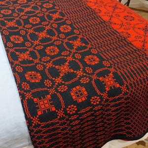 Op de afbeelding: Een zwart-rood patroon deken met een geometrisch ontwerp. De deken is over een bed gevouwen en toont het ingewikkelde geweven patroon. Het kleurenschema is een opvallend contrast van zwart en rood, waardoor een visueel aantrekkelijk textiel ontstaat.
