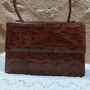 May include: A vintage brown handbag with a snakeskin pattern. The rectangular bag has a curved handle and a flap closure. The bag is made of a textured material and sits on a white lace cloth.