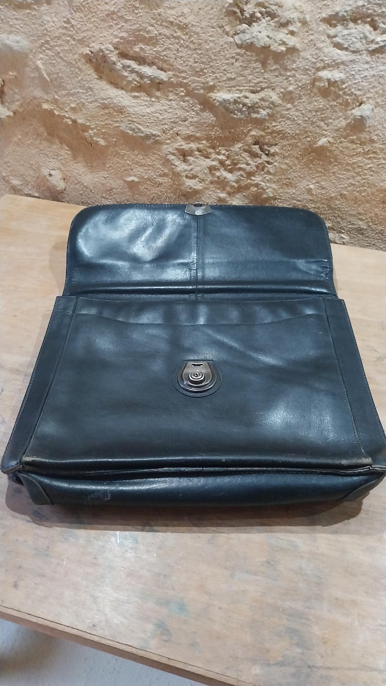 Puede incluir: Malet&iacute;n de cuero negro abierto con un cierre met&aacute;lico. El malet&iacute;n tiene forma rectangular con una solapa que se abre para revelar el interior. El cuero parece desgastado, lo que sugiere edad y uso. El fondo es una pared texturizada de color claro.