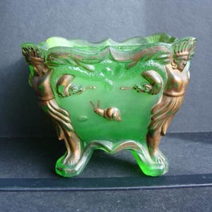 May include: A green glass planter with bronze-coloured accents. The planter features a sculpted design with figures on the sides and a snail motif. The planter is supported by three feet.