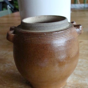 May include: A brown ceramic crock with two handles. The crock has a wide mouth and a rounded body.
