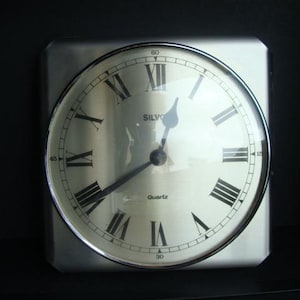 May include: A square, silver-toned quartz clock with a circular face. The clock face features black Roman numerals and hands, with the word "SILVO" and "Quartz" printed on the face. The clock is set against a black background.