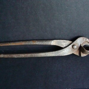 Pode incluir: Uma ferramenta de corte de metal vintage com um acabamento enferrujado. A ferramenta tem duas alças longas e uma cabeça arredondada com uma aresta de corte curva. Os alicates são projetados para cortar ou agarrar.