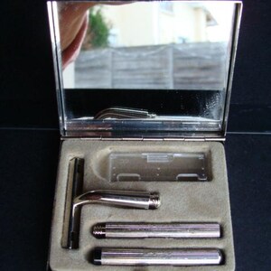 Peut inclure: Un nécessaire de rasage argenté vintage avec miroir intégré. Le kit comprend un rasoir de sûreté, deux manches cylindriques et un porte-lame en plastique transparent, le tout niché dans un étui doublé de velours beige. L'étui est ouvert, révélant le contenu.