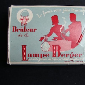 Peut inclure: Une boîte bleue et blanche avec du texte rouge pour un brûleur à huile Lampe Berger. Le texte indique "La fumée vous gêne Madame ? Le Brûleur de la Lampe Berger absorbe fumée de tabac et odeurs désagréables. Made in France."