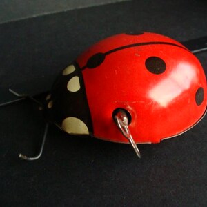 May include: A vintage wind-up toy ladybug. The ladybug is primarily red with black spots and a black head with white spots. It has metal antennae and a winding key.