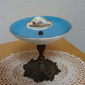 Può includere: Un piatto in porcellana blu e bianco con un bordo dorato e un dipinto di un cherubino. Il piatto è su un piedistallo in bronzo con una base decorativa.