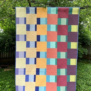 Może przedstawiać: Kolorowa kołdra z geometrycznym wzorem w odcieniach niebieskiego, żółtego, zielonego, czerwonego i fioletowego. Kołdra jest powieszona na sznurze do suszenia w ogrodzie.