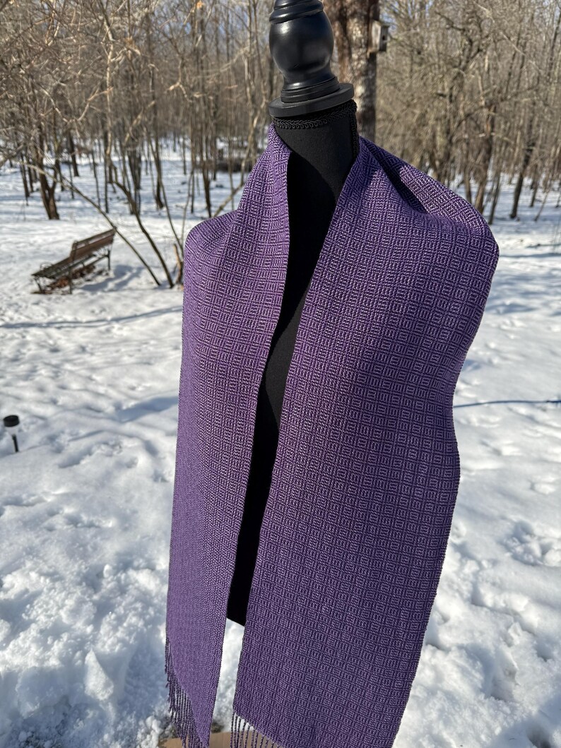 Puede incluir: Una bufanda morada con un patr&oacute;n geom&eacute;trico est&aacute; drapeada sobre un maniqu&iacute; negro. La bufanda tiene un dise&ntilde;o de tejido de diamantes y flecos en los extremos. El fondo es una escena nevada al aire libre con &aacute;rboles.