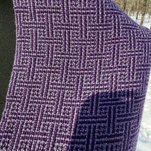 Puede incluir: Primer plano de un textil tejido con un patr&oacute;n geom&eacute;trico en tonos morados y blancos. La tela tiene una apariencia texturizada, con un dise&ntilde;o cuadrado repetitivo. El textil est&aacute; drapeado, mostrando el intrincado tejido y las variaciones de color.