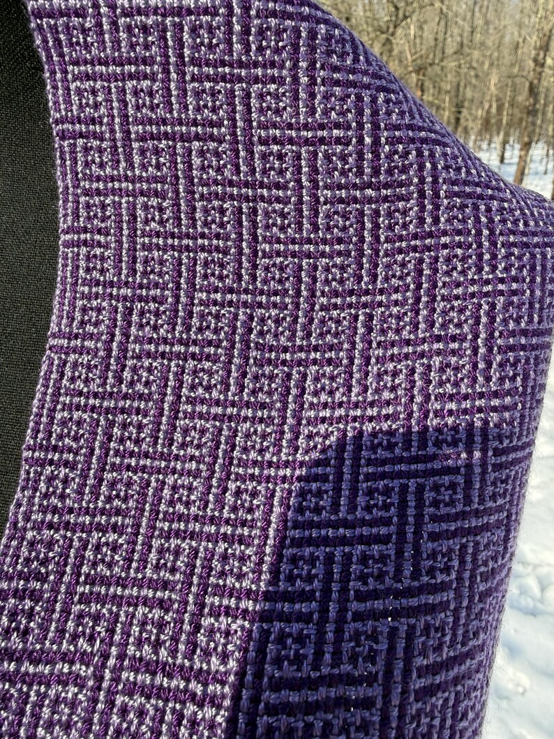 Puede incluir: Primer plano de un textil tejido con un patr&oacute;n geom&eacute;trico en tonos morados y blancos. La tela tiene una apariencia texturizada, con un dise&ntilde;o cuadrado repetitivo. El textil est&aacute; drapeado, mostrando el intrincado tejido y las variaciones de color.