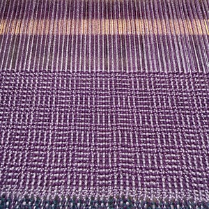Puede incluir: Primer plano de un textil tejido en tonos morado, blanco y turquesa. La tela presenta un patr&oacute;n geom&eacute;trico con flecos en la parte inferior. El proceso de tejido es visible, mostrando los hilos y la estructura del telar. El textil es probablemente una bufanda o un art&iacute;culo similar.
