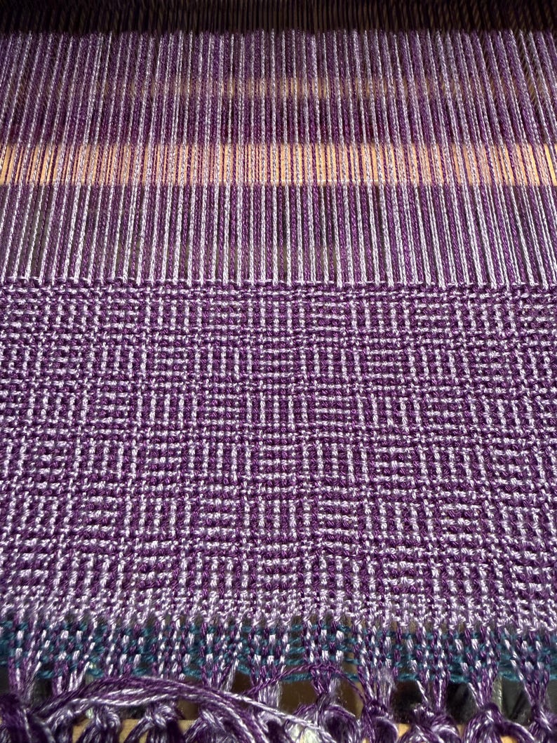 Puede incluir: Primer plano de un textil tejido en tonos morado, blanco y turquesa. La tela presenta un patr&oacute;n geom&eacute;trico con flecos en la parte inferior. El proceso de tejido es visible, mostrando los hilos y la estructura del telar. El textil es probablemente una bufanda o un art&iacute;culo similar.